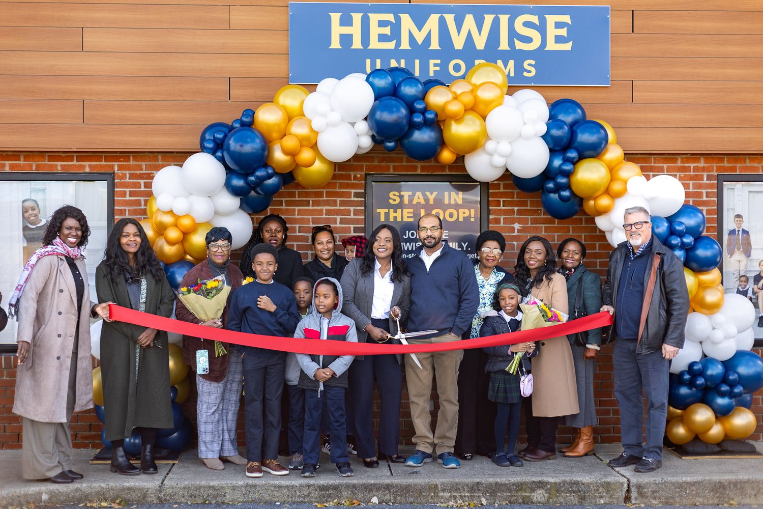 A group gathers for a ribbon cutting ceremony in front of Hemwise with blue and gold balloon decorations.