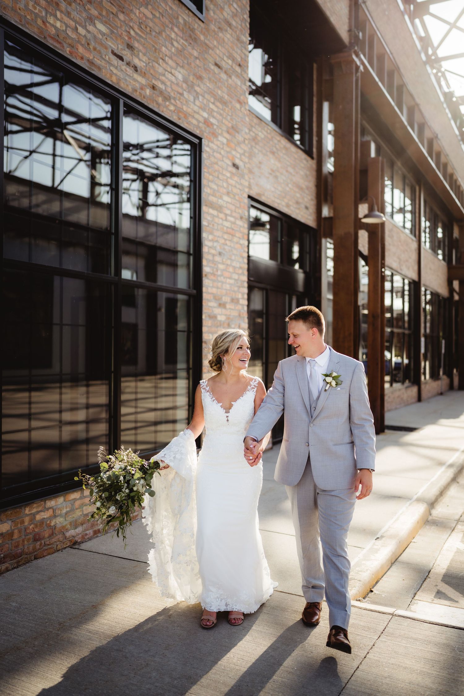 Bride and groom walk hand-in-hand outside an industrial brick building on their wedding day.