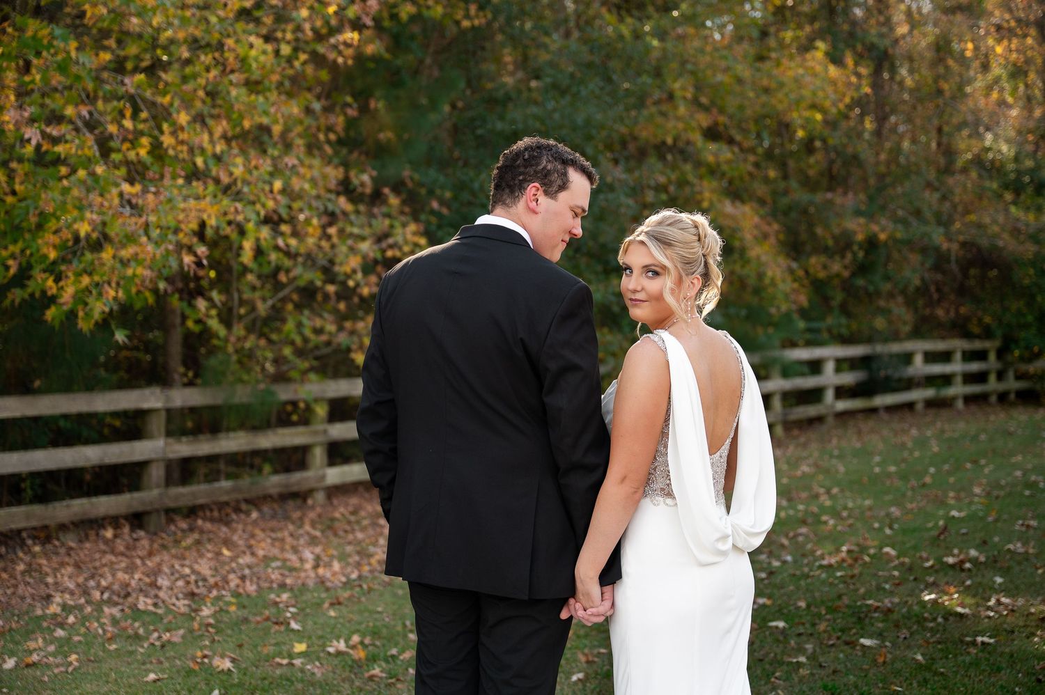 Autumn wedding portrait with couple standing on grass near wooden fence.
