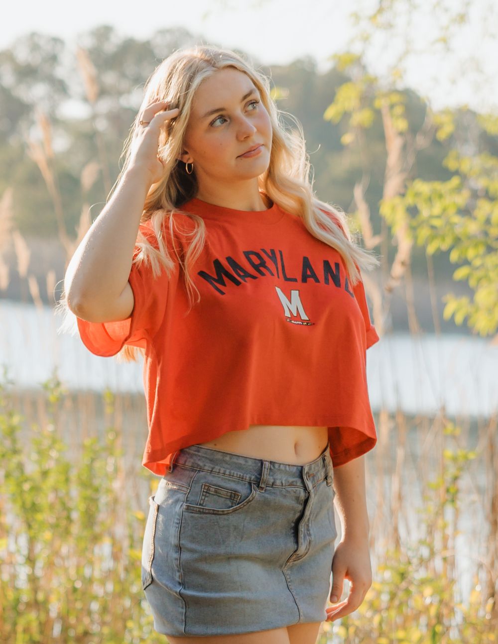 Blonde woman wearing a red Maryland crop top and denim skirt posing outdoors near water.