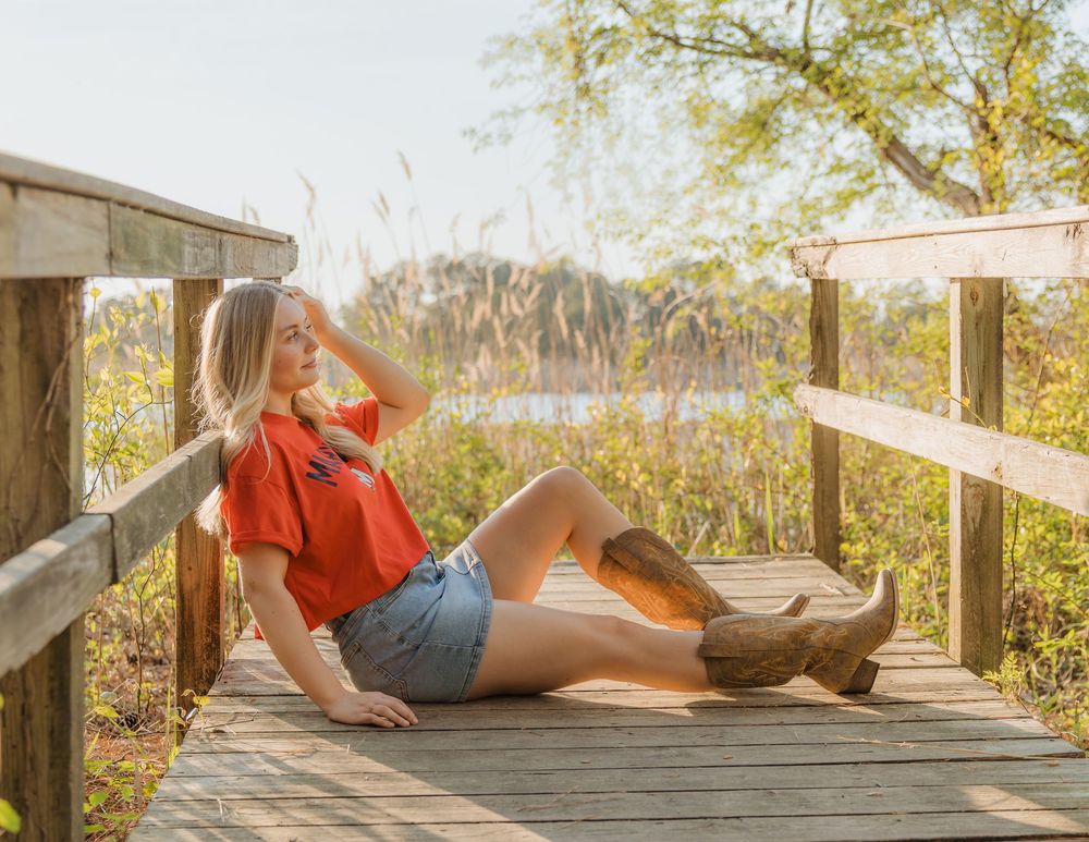 Woman in red top, denim shorts, and cowboy boots relaxing on a sunny wooden dock surrounded by nature.