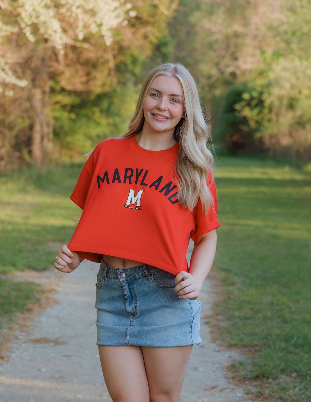 Young woman smiling in a red Maryland University crop top and denim skirt on a scenic outdoor path.