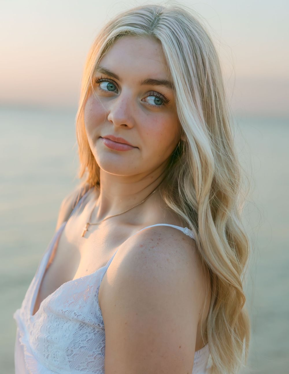 Young blonde woman with blue eyes wearing a white lace dress poses near the ocean at golden hour.