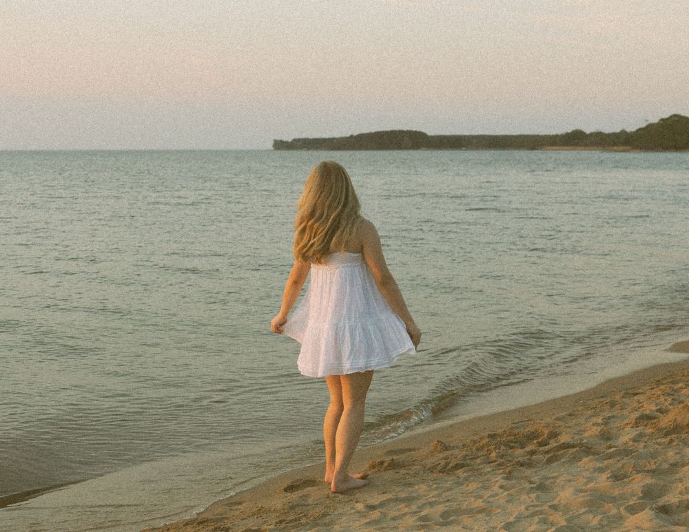 A young blonde girl in a white dress walks barefoot on a sandy beach at sunset with calm ocean waters.