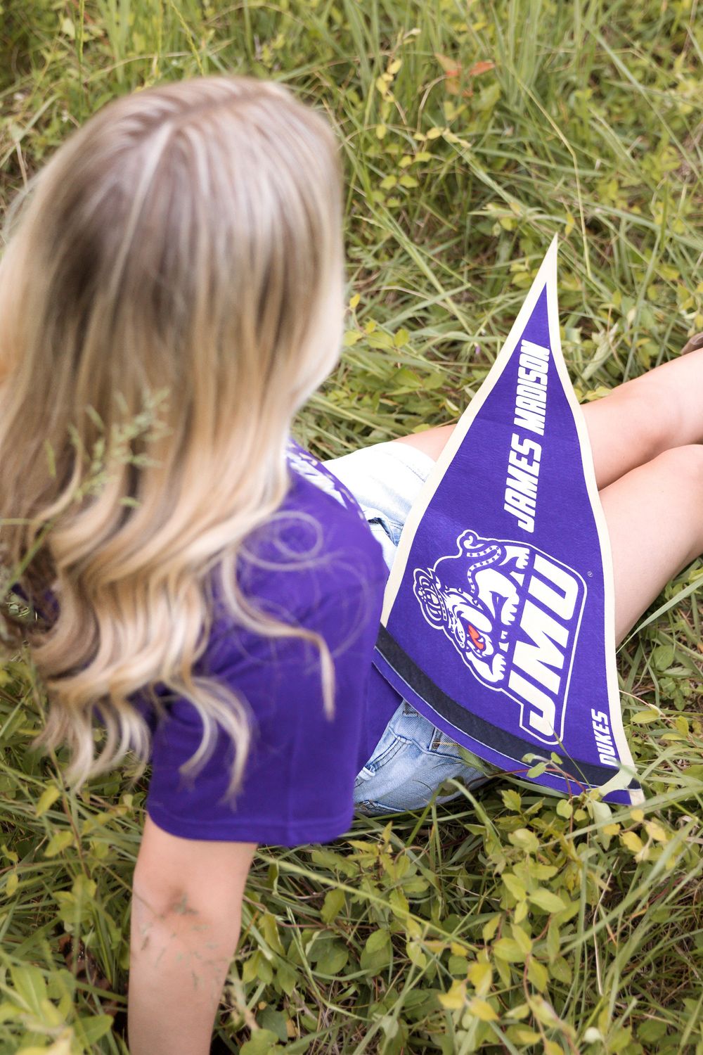 Blonde girl sitting in grass wearing purple JMU James Madison University t-shirt holding a JMU pennant flag.
