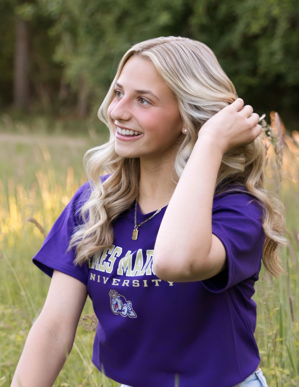 Smiling blonde woman wearing a purple university t-shirt poses outdoors in a field with tall grass.