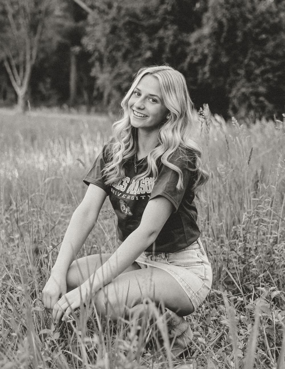 Black and white photo of a smiling young blonde woman sitting in a field of tall grass wearing shorts and a t-shirt.