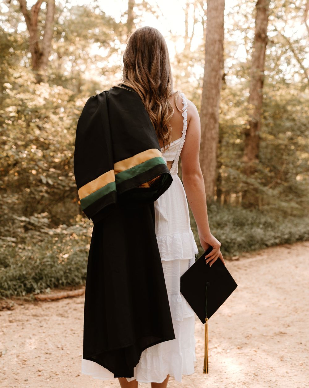 A couple in wedding attire walks hand in hand down a sunlit forest path during golden hour.