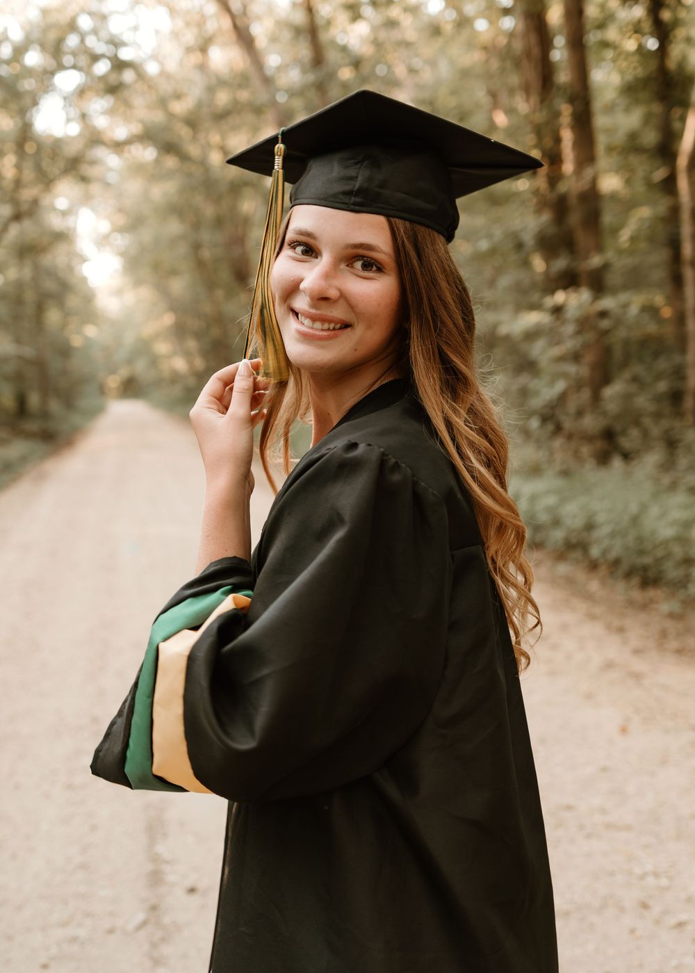 A graduate in cap and gown poses on a wooded path during a sunny outdoor graduation photoshoot.