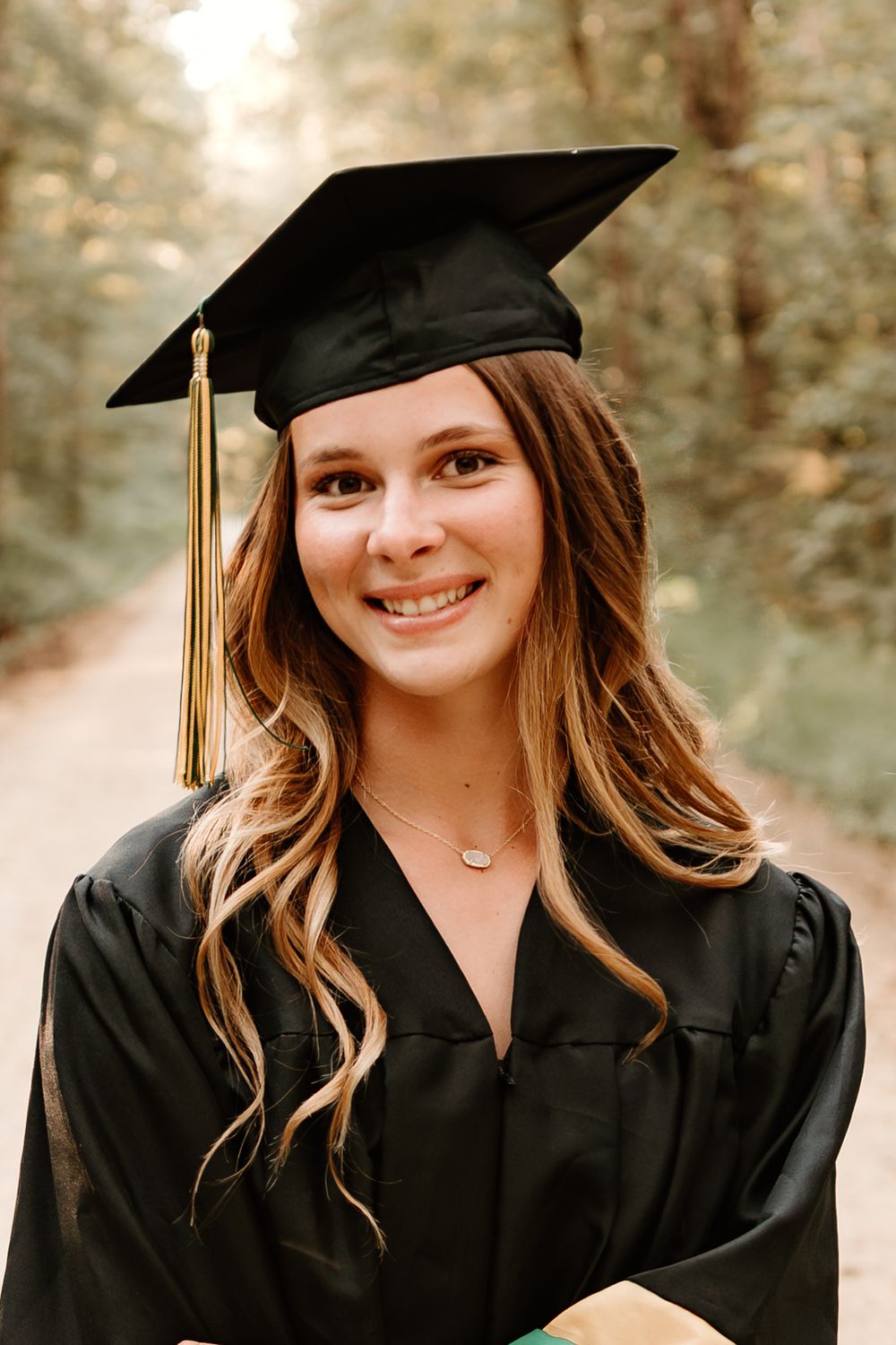 Graduate in black cap and gown with gold tassel smiling outdoors with soft natural lighting in background.