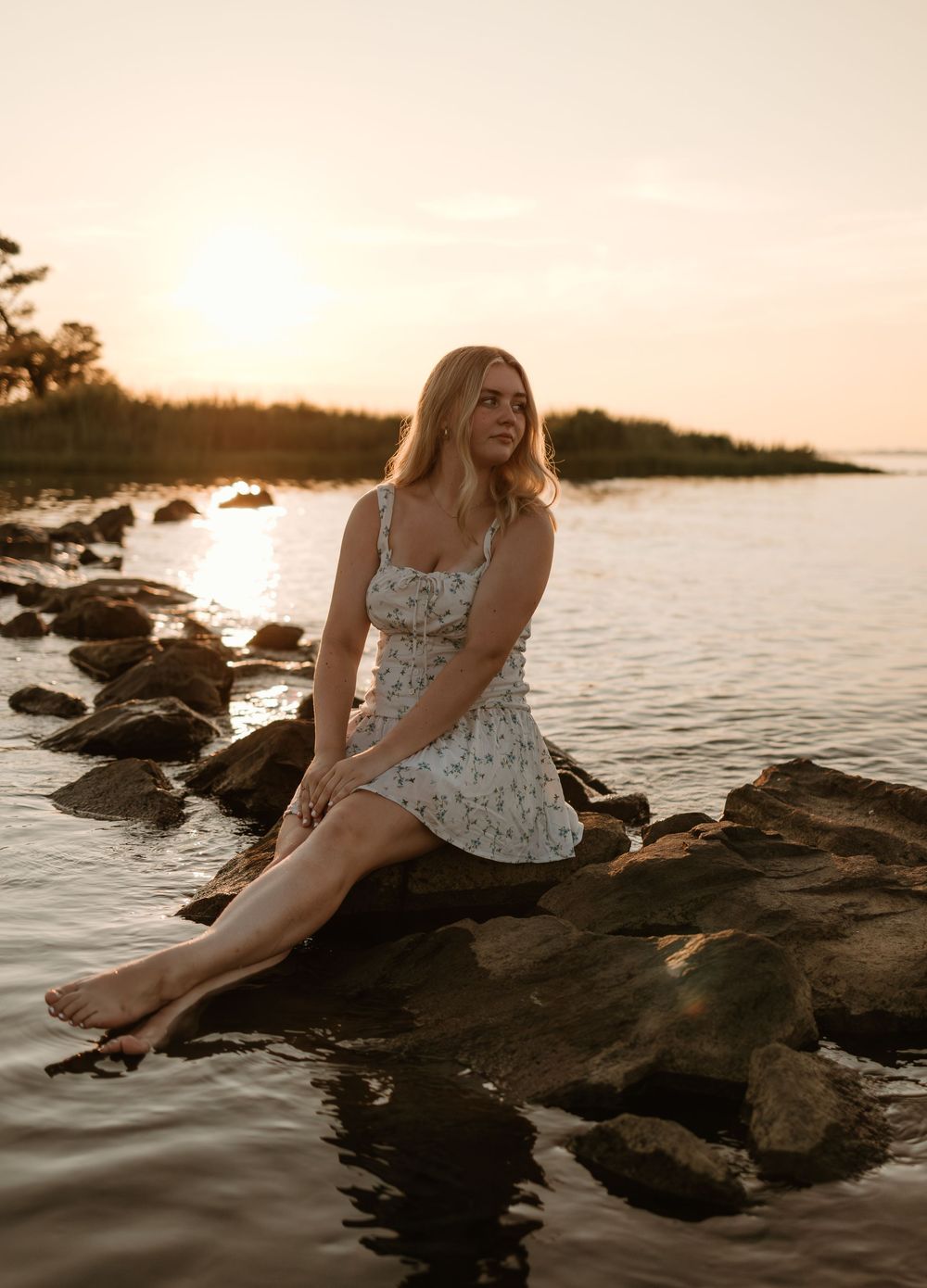 Person in a floral dress poses on rocky shoreline at sunset as golden light reflects off calm ocean waters.