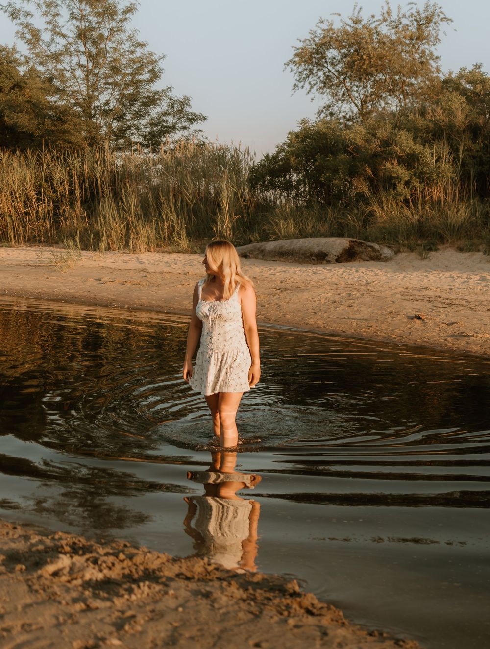 Person in a white dress wades in shallow water at sunset creating rippling reflections on a sandy beach.