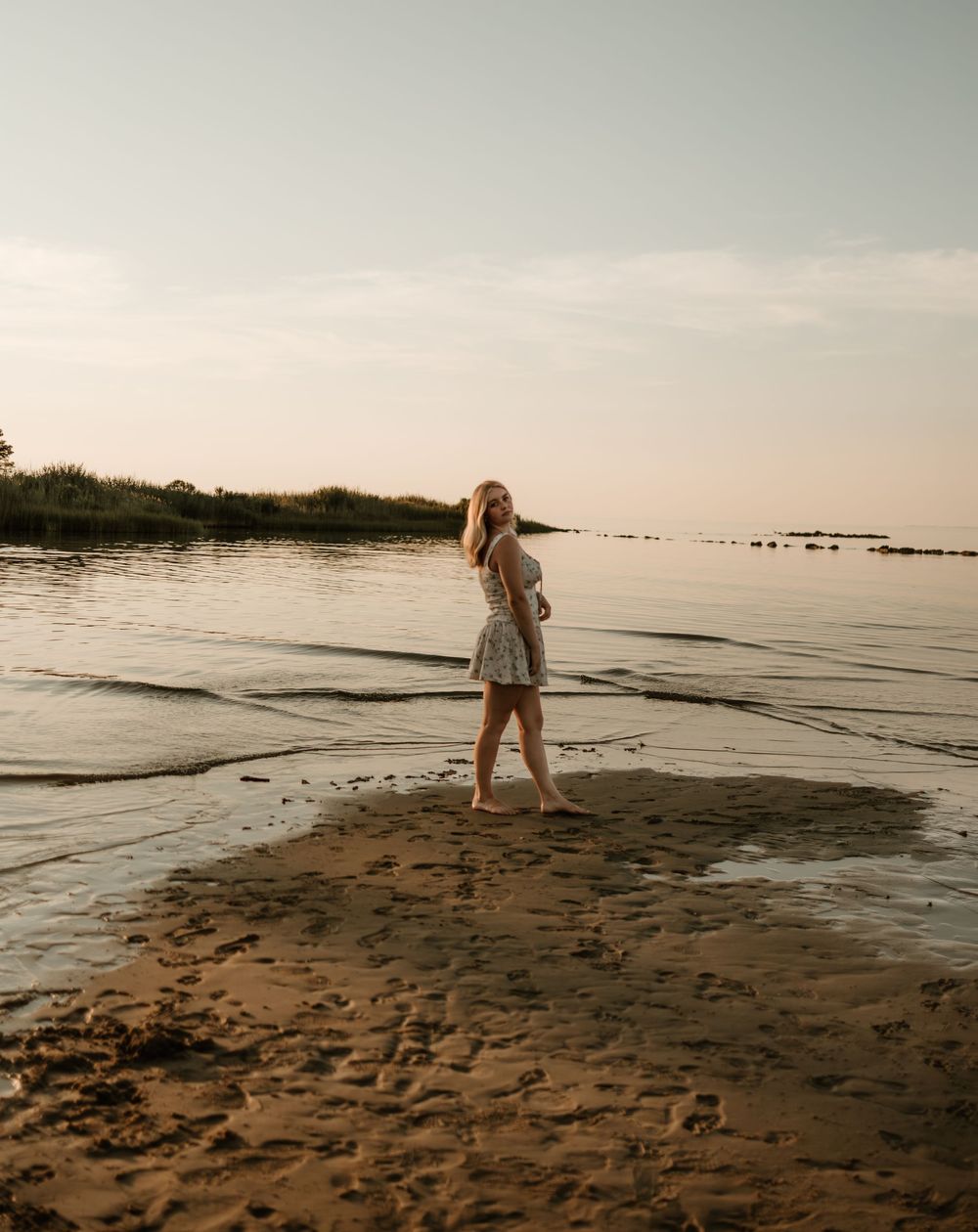 A person in casual attire explores a tranquil beach shoreline at sunset while the gentle waves lap at the sandy shore.