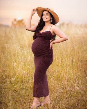 Maternity photoshoot in a golden field at sunset with a brown dress and wide-brimmed hat.