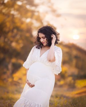 A woman in a flowing white dress poses in a golden autumn field at sunset, creating an ethereal maternity portrait.