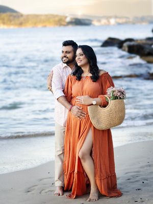 A couple in matching orange dresses poses together on a rocky beach at sunset with a woven basket.