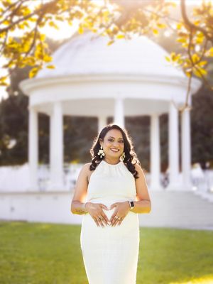 A person in a white dress poses outdoors near white gazebos during golden hour sunlight.