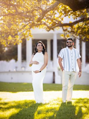 Couple poses under golden autumn tree leaves during maternity photoshoot on grassy lawn with white building in background.
