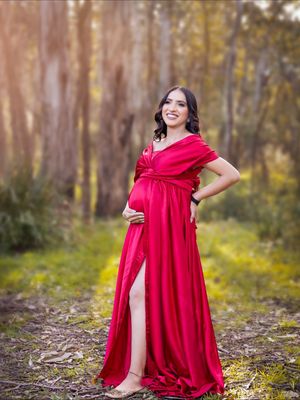 A woman in a flowing red dress poses gracefully in a wooded area with dappled sunlight and fallen leaves.