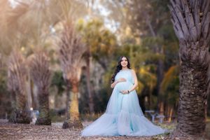 A person in a flowing light blue gown poses elegantly among autumn trees and fallen leaves in an outdoor setting.