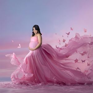 A flowing pink maternity gown billows dramatically against a sunset sky with floating butterflies creating a dreamy scene.