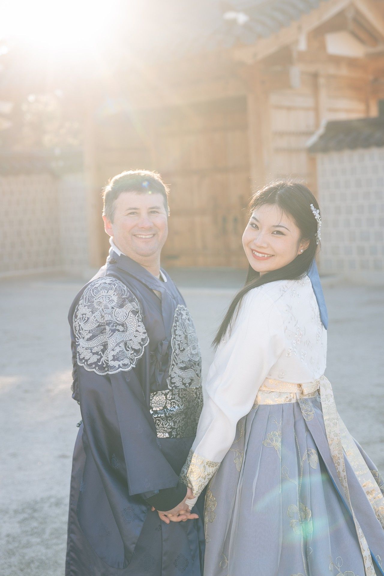 A couple in traditional Korean hanbok attire poses together in a sunlit courtyard.