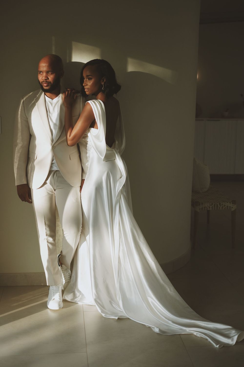 Black couple in white wedding attire posing dramatically in sunlit modern interior space.