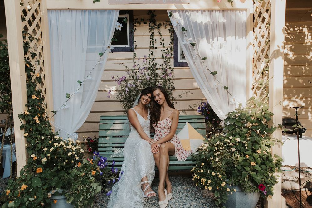 Two women in wedding attire sit together under a floral garden arbor with white curtains.