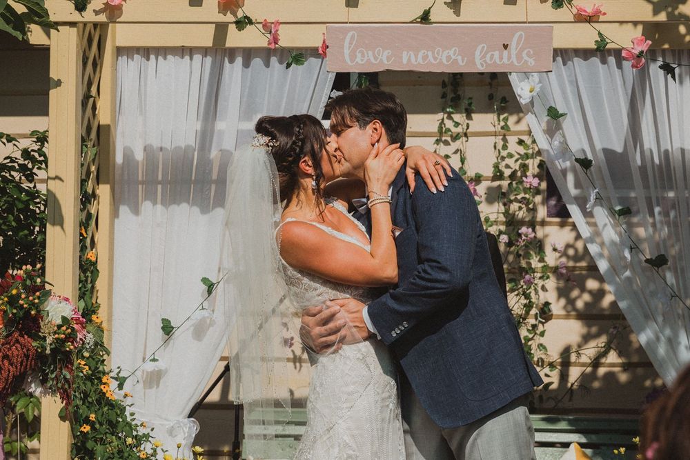 Bride and groom share their first kiss under a floral arch with love never fails sign.