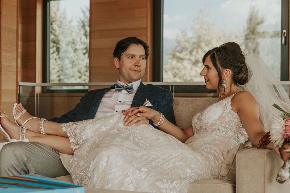Bride and groom sit together on a couch holding hands at their mountain wedding venue.