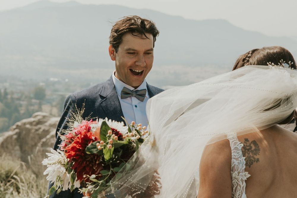 Joyful groom laughs while holding bridal bouquet of red flowers at outdoor mountain wedding.