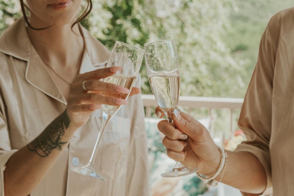 Two women clinking champagne flutes in a celebratory toast at an outdoor wedding event.