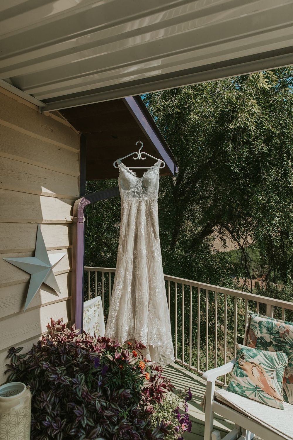 Lace wedding dress hanging on a hanger on a sunny outdoor porch with green trees in the background.