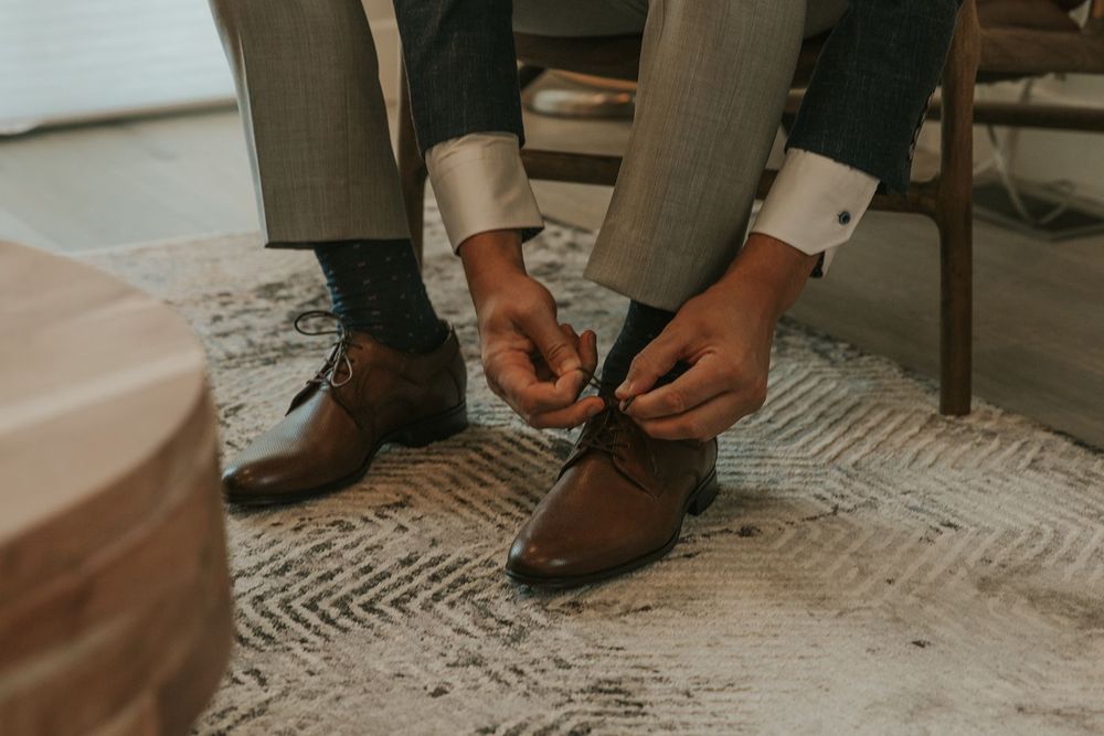 Groom tying brown leather dress shoes on a patterned rug while preparing for his wedding day.