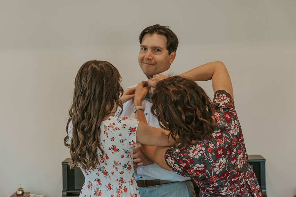 Two women helping a smiling groom adjust his boutonniere while getting ready for his wedding.