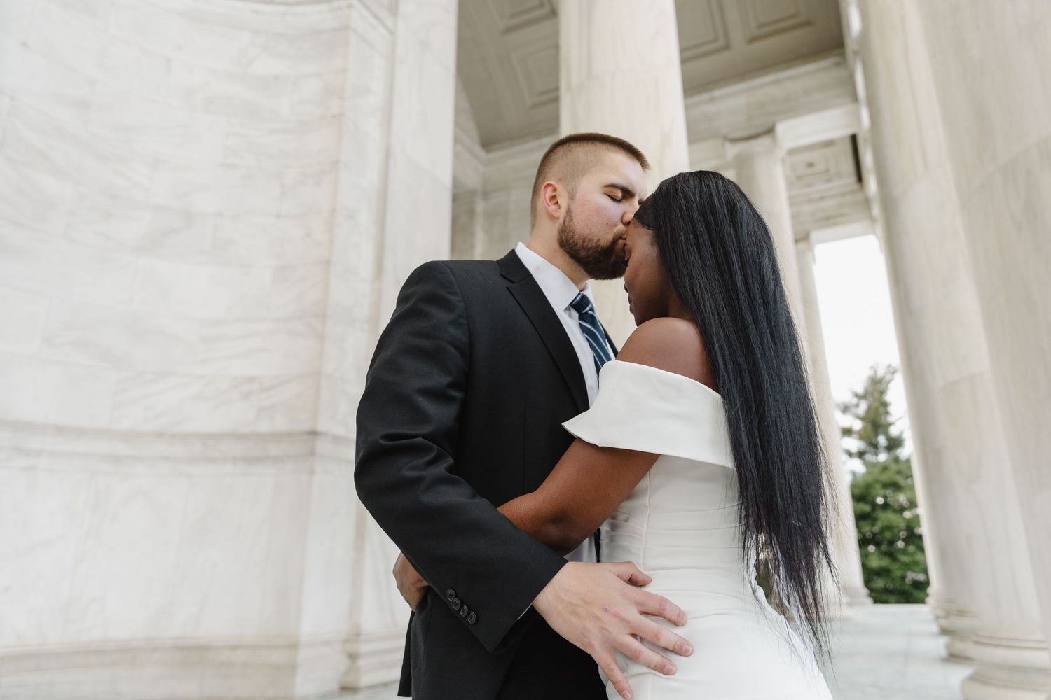 A joyful wedding couple shares a romantic moment among white columns while holding a red rose bouquet.