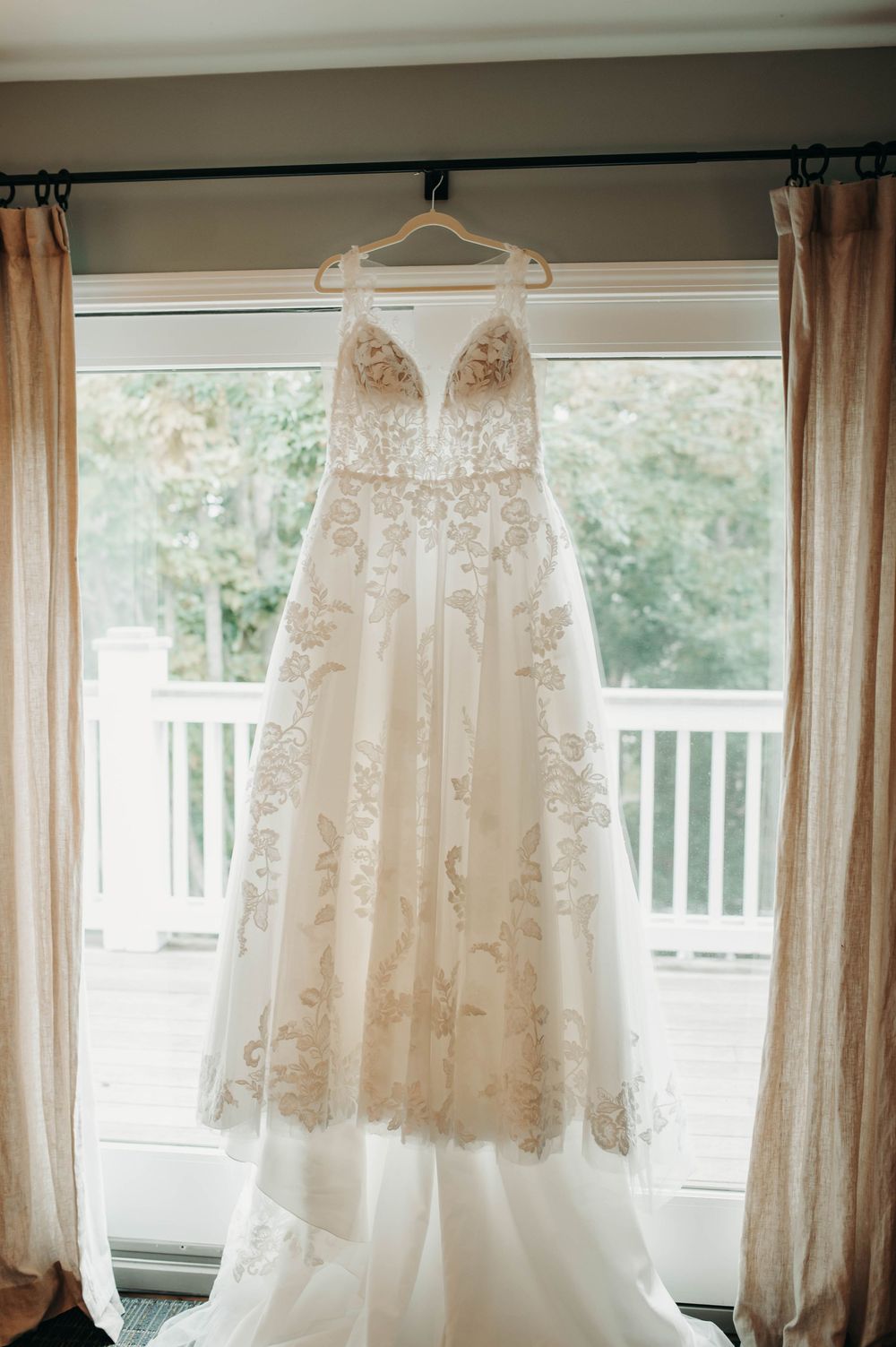 Elegant lace A-line wedding dress with floral embroidery hanging in window with natural light.