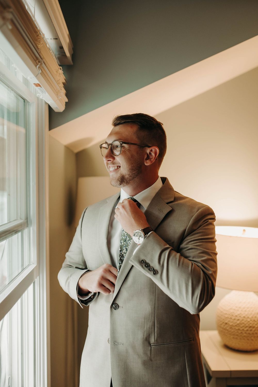 Smiling groom in tan suit adjusting floral tie near window in warm indoor light.