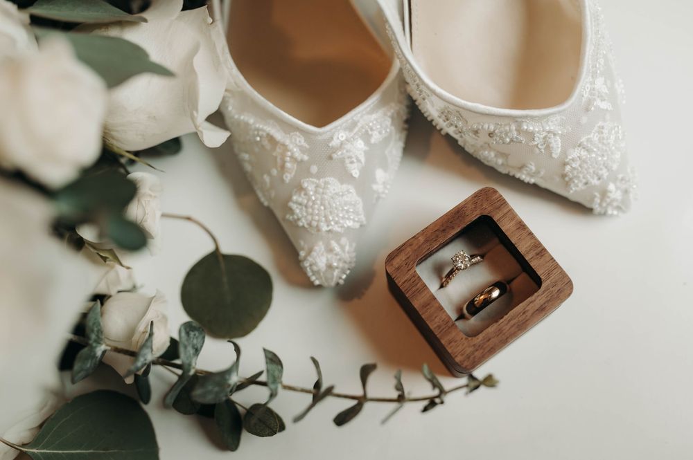 White lace bridal heels with wedding rings in wooden box and eucalyptus sprigs.