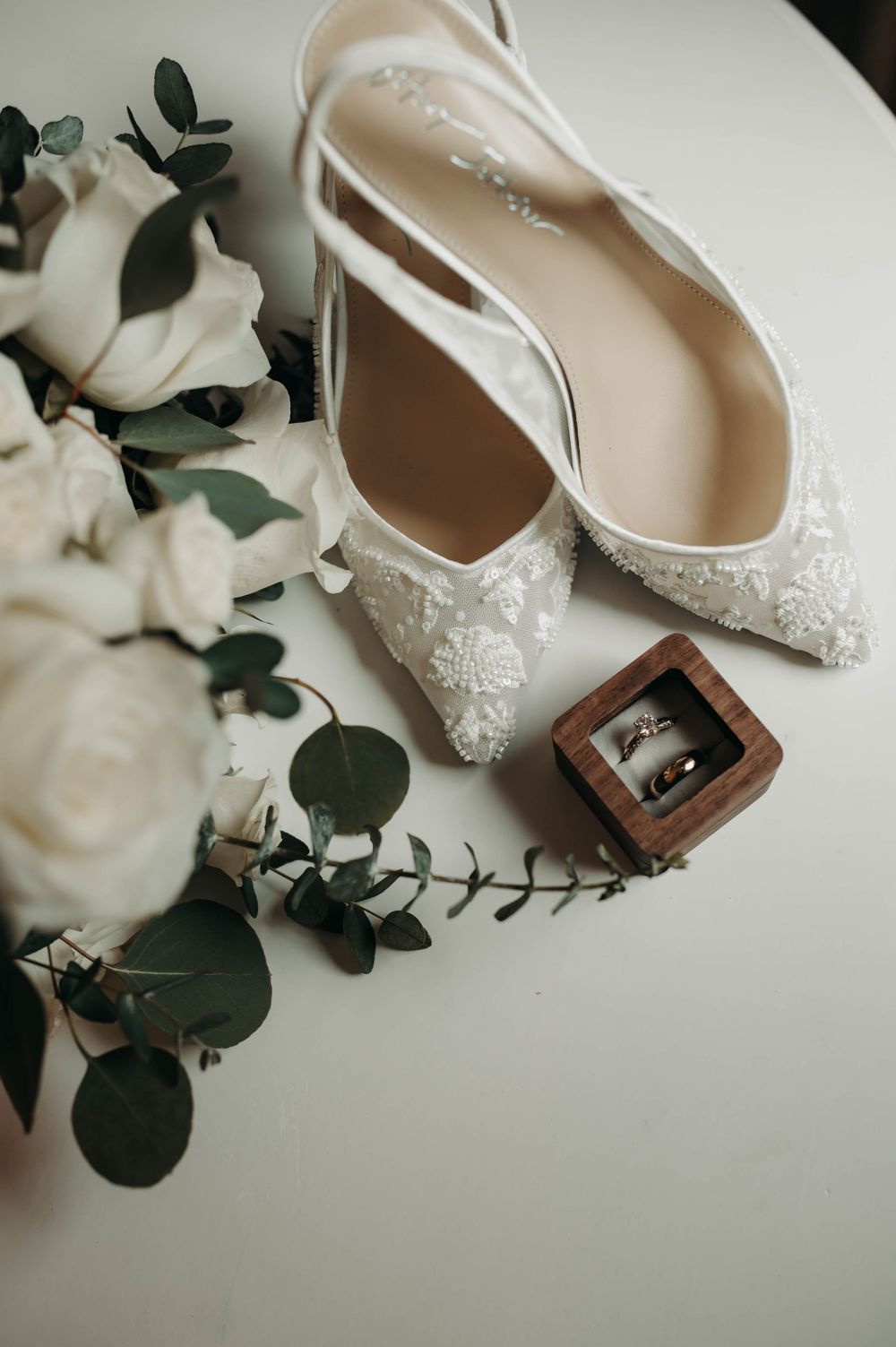 White lace bridal heels beside white rose bouquet and wedding rings in wooden box.
