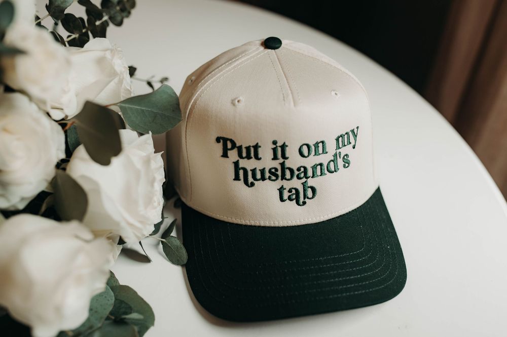 Novelty cap reading Put it on my husbands tab beside white wedding flowers.