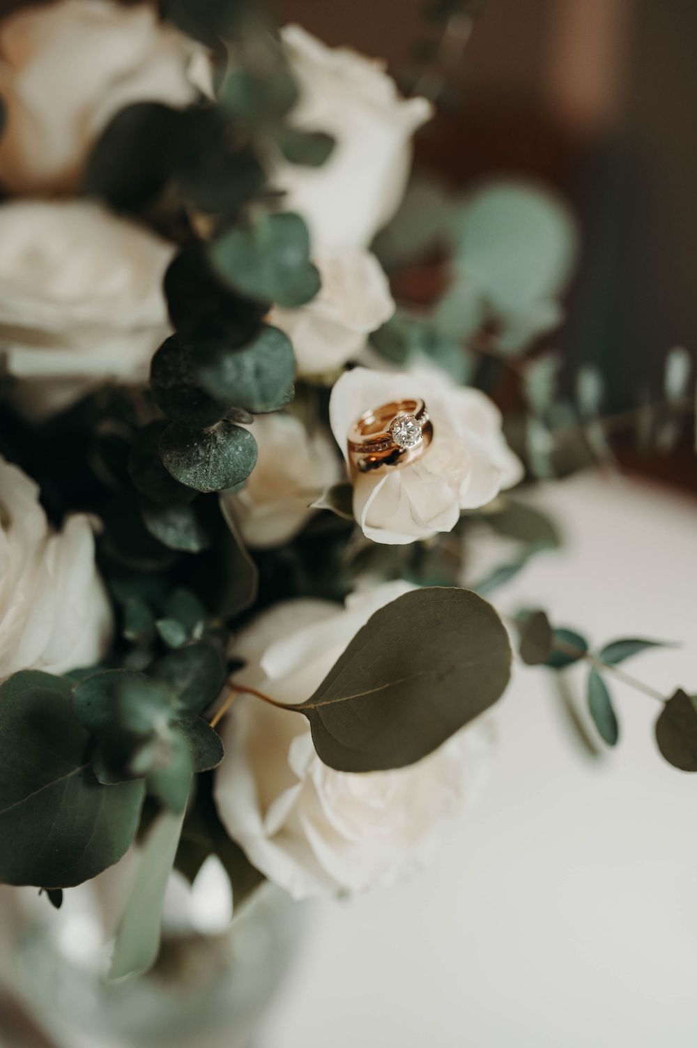 Gold wedding rings resting on white roses and eucalyptus in bridal bouquet.