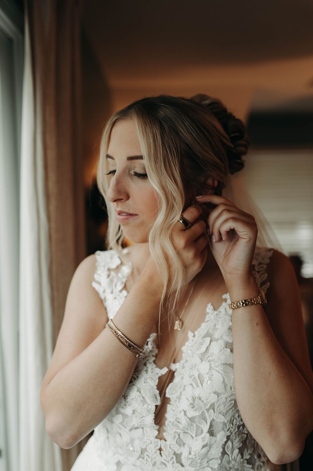 Bride in floral lace wedding gown adjusting earring near window with elegant updo hairstyle.