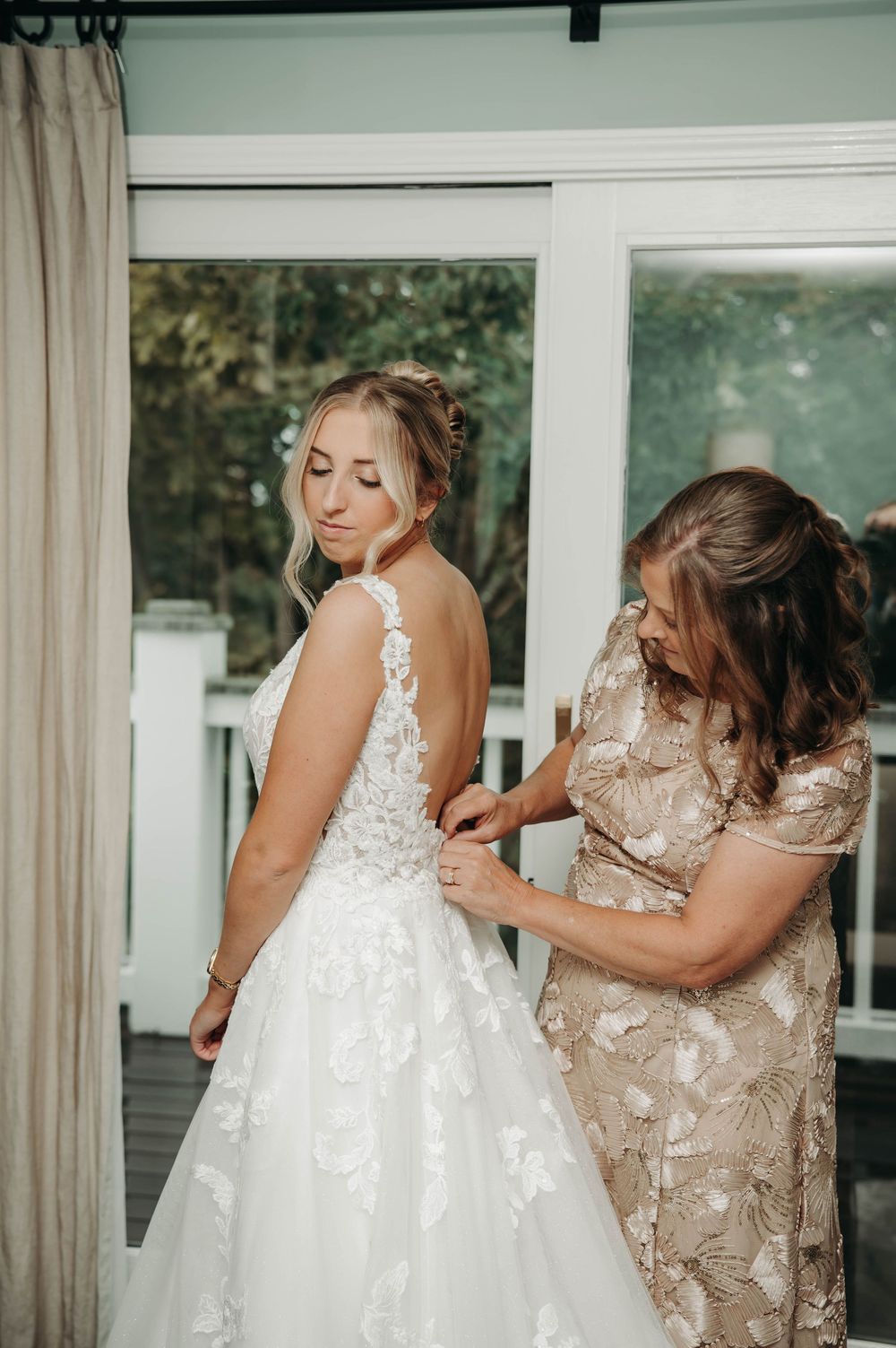 Mother helps bride fasten the back of her lace wedding gown near a bright window.