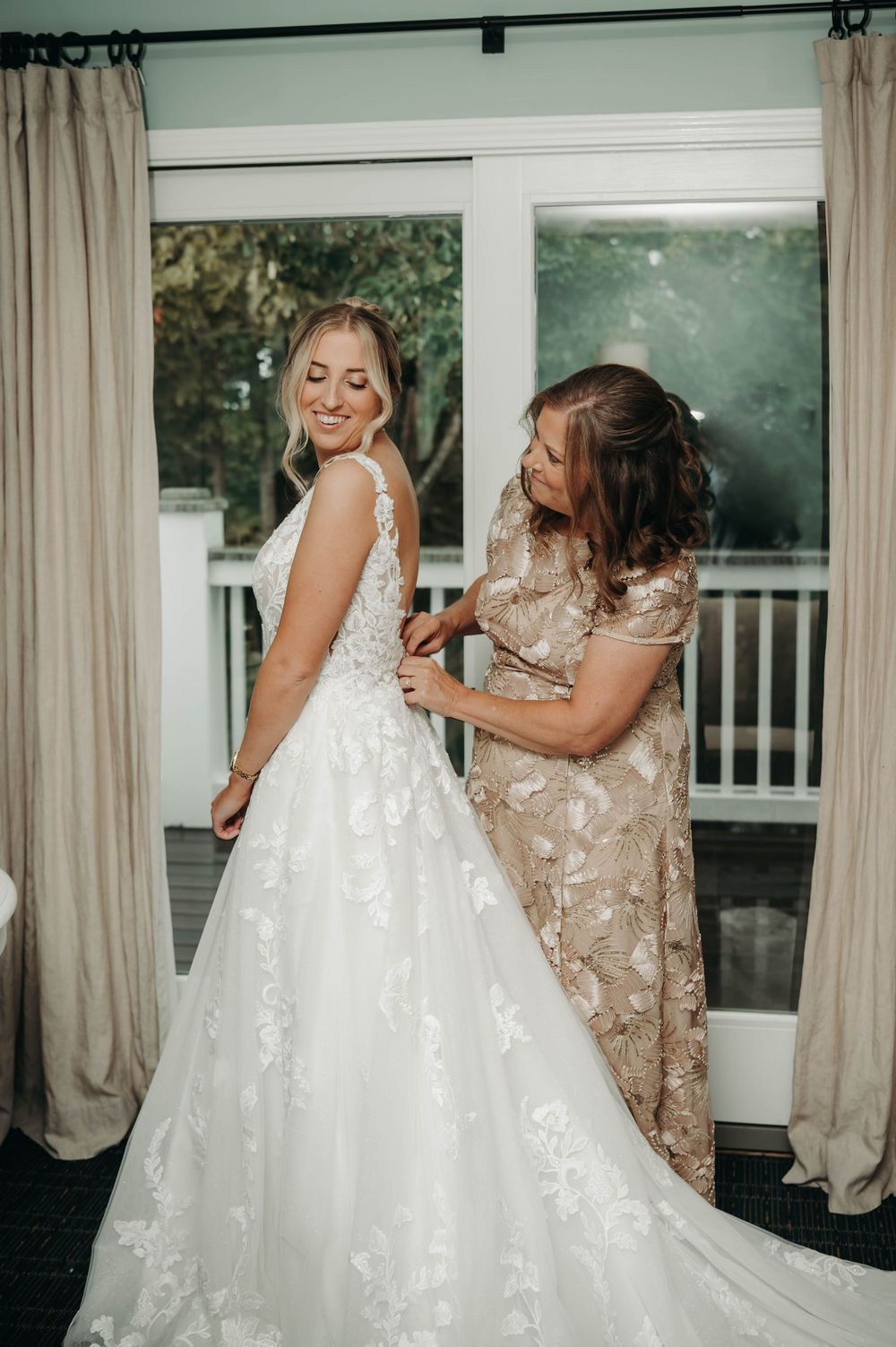 Smiling bride gets her lace wedding dress fastened by her mother before the ceremony.
