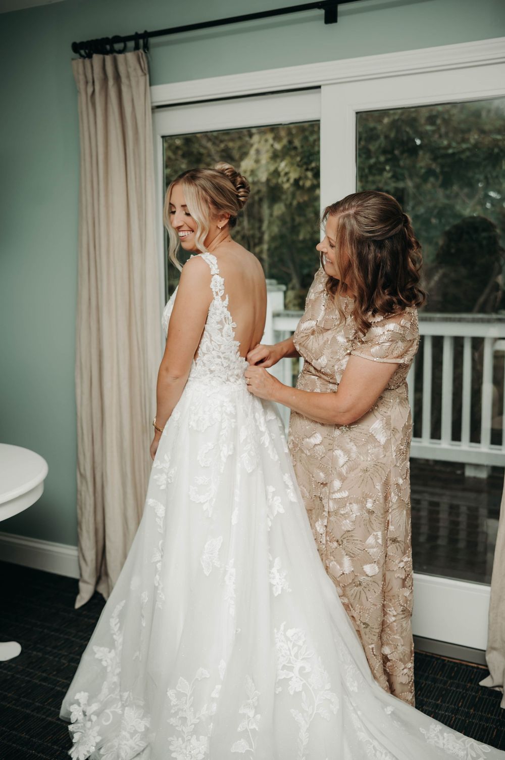 Laughing bride has her elegant lace wedding dress buttoned by her mother before ceremony.