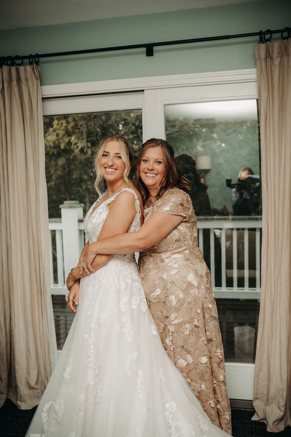 Bride in white lace gown poses with mother in gold floral dress during wedding day preparations.