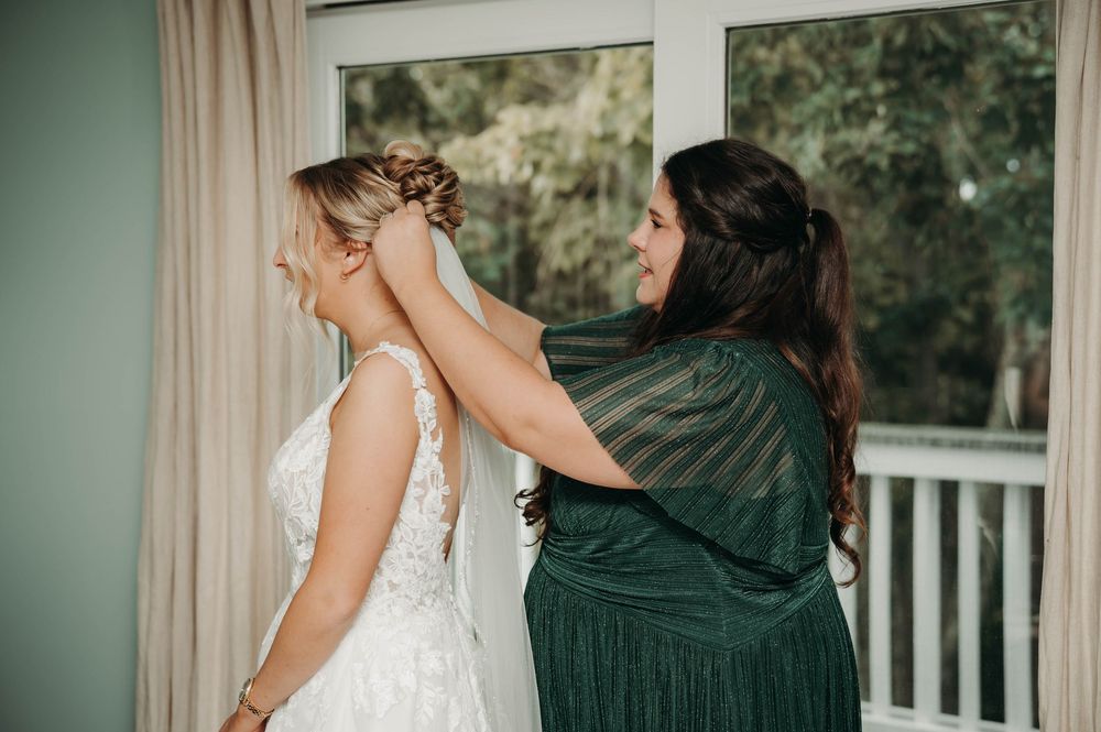 Bridesmaid in green dress carefully adjusts bride's updo hairstyle before wedding ceremony.