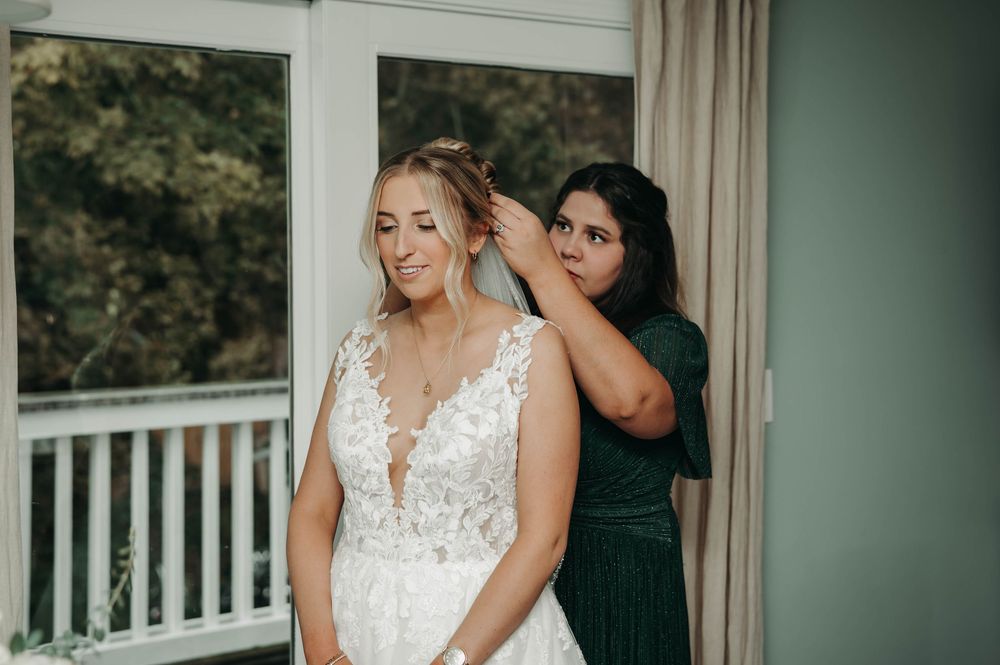 Smiling bride in lace wedding gown has her hair styled by bridesmaid in emerald green dress.
