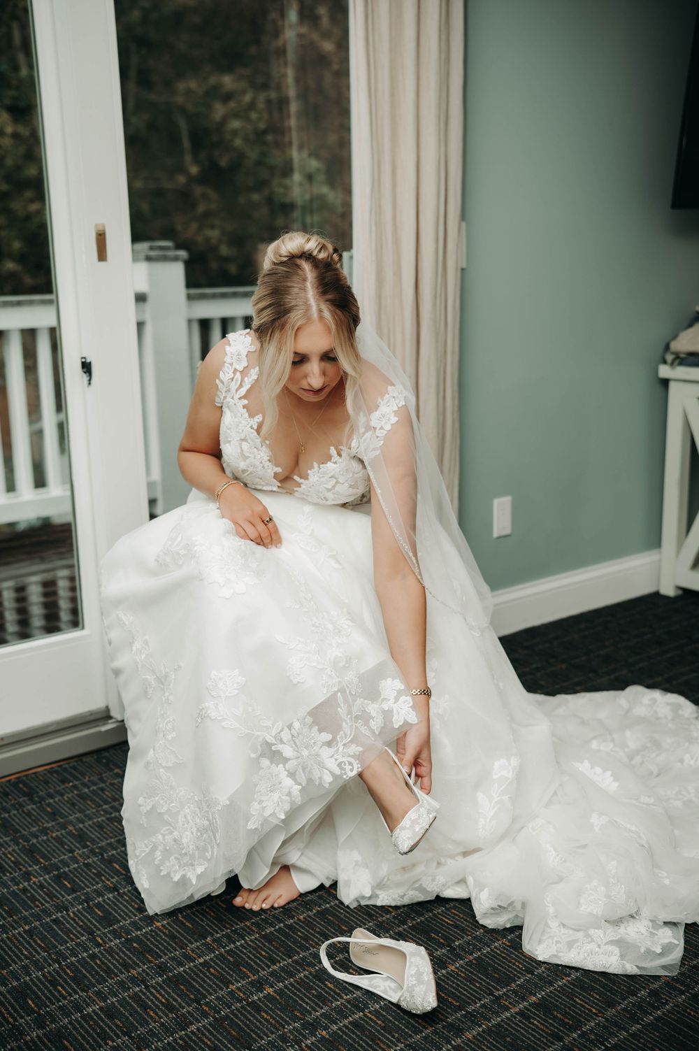 Bride in lace ball gown sits on floor putting on white heels before her wedding ceremony.